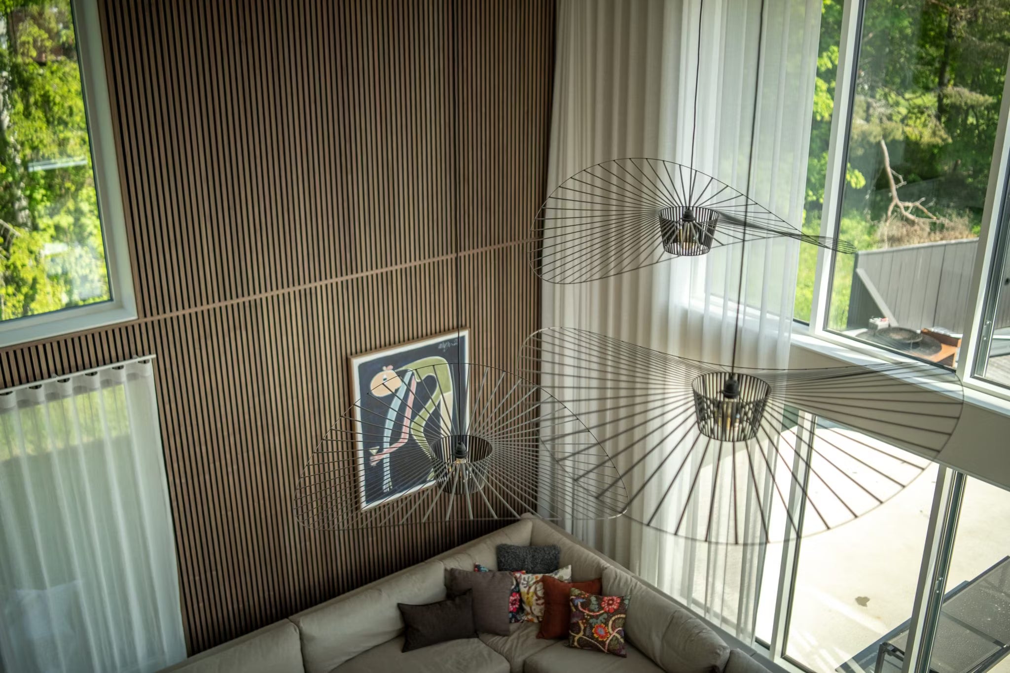 Modern living room with Nordic wood Akupanel walls, large windows, abstract acoustic panel art, a sectional sofa, and minimalist wireframe light fixtures, viewed from above.