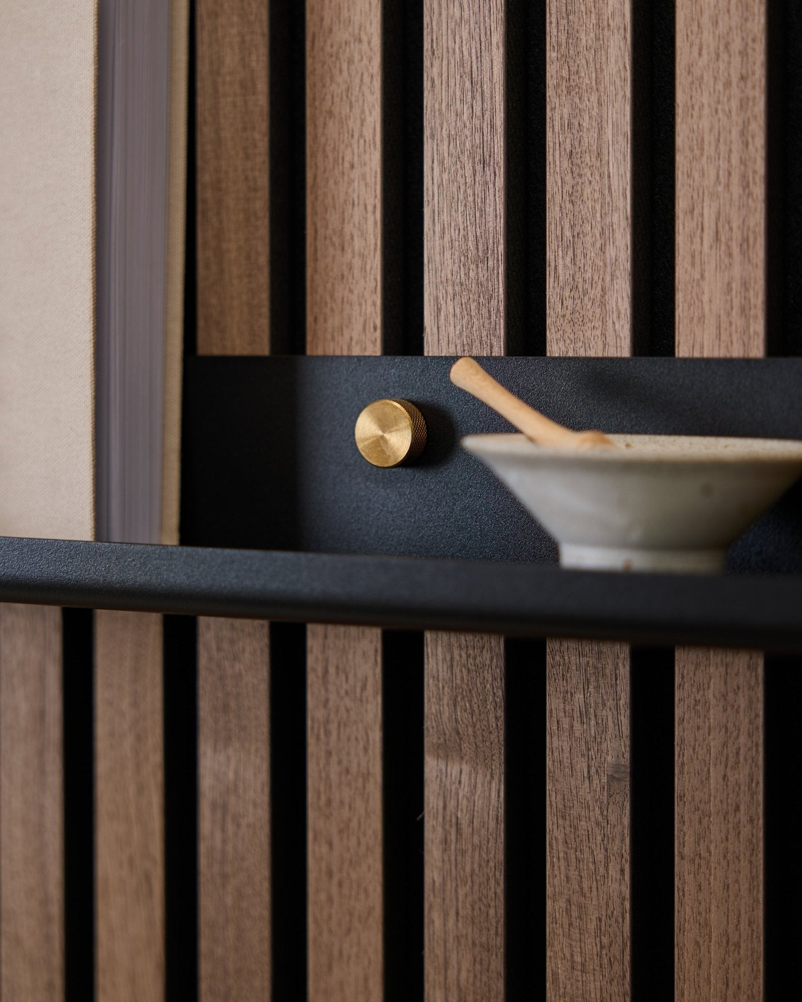 Close-up of a 115.9 cm black WoodUpp Norr Narrow Shelf mounted on a Nordic acoustic slatwall panel with brass thumbscrews, gold knob, and ceramic bowl with spoon.