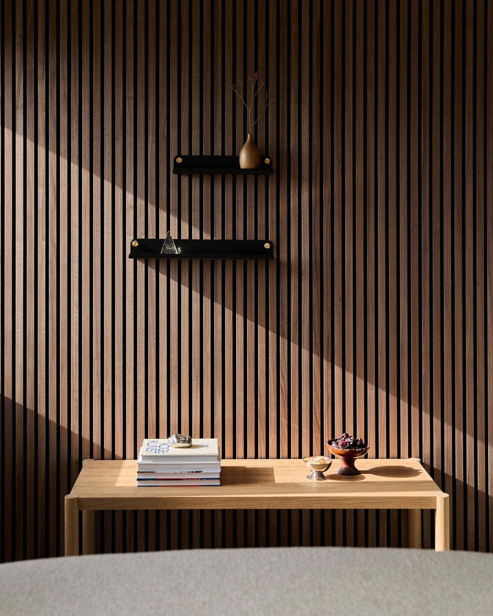 A minimalist wooden table with books and bowls sits by a Nordic slatwall acoustic panel, highlighting the WoodUpp Norr Narrow Shelf in black (51.9 cm); sunlit shadows above.