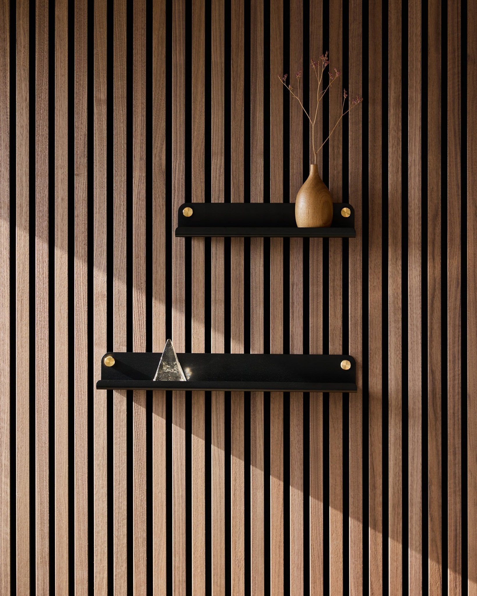 Two black WoodUpp Norr Narrow shelves (35.9 cm) mounted on a Nordic acoustic slatwall panel; top shelf with vase of dried branches, bottom with glass pyramid.