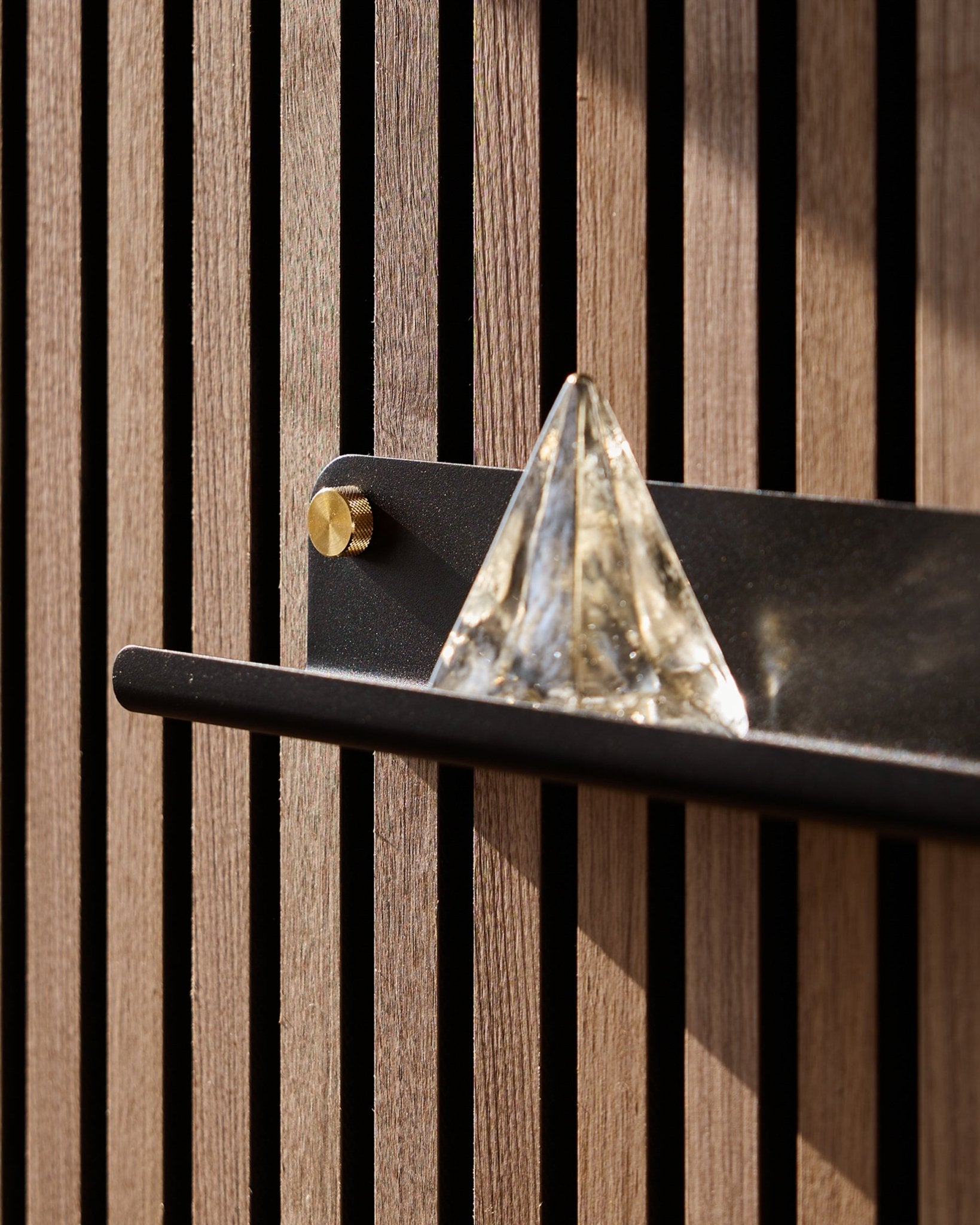 A glass pyramid sits on the WoodUpp Norr Narrow Shelf in black (51.9 cm), mounted on a vertical acoustic panel and highlighted by sunlight.