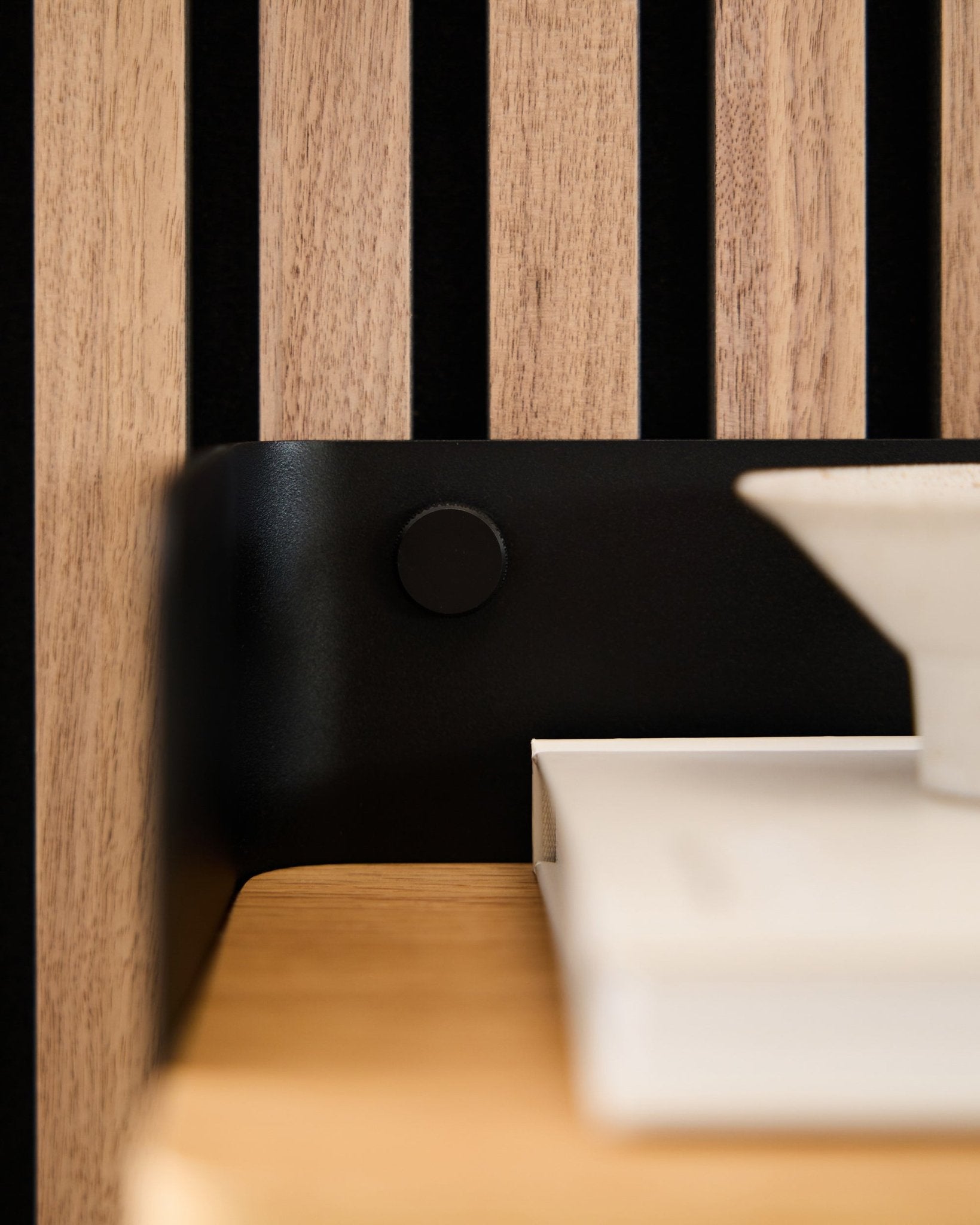 Close-up of a WoodUpp Akupanel acoustic wood slatwall panel with black Finger screws securing a black bracket, alongside white books and a bowl.