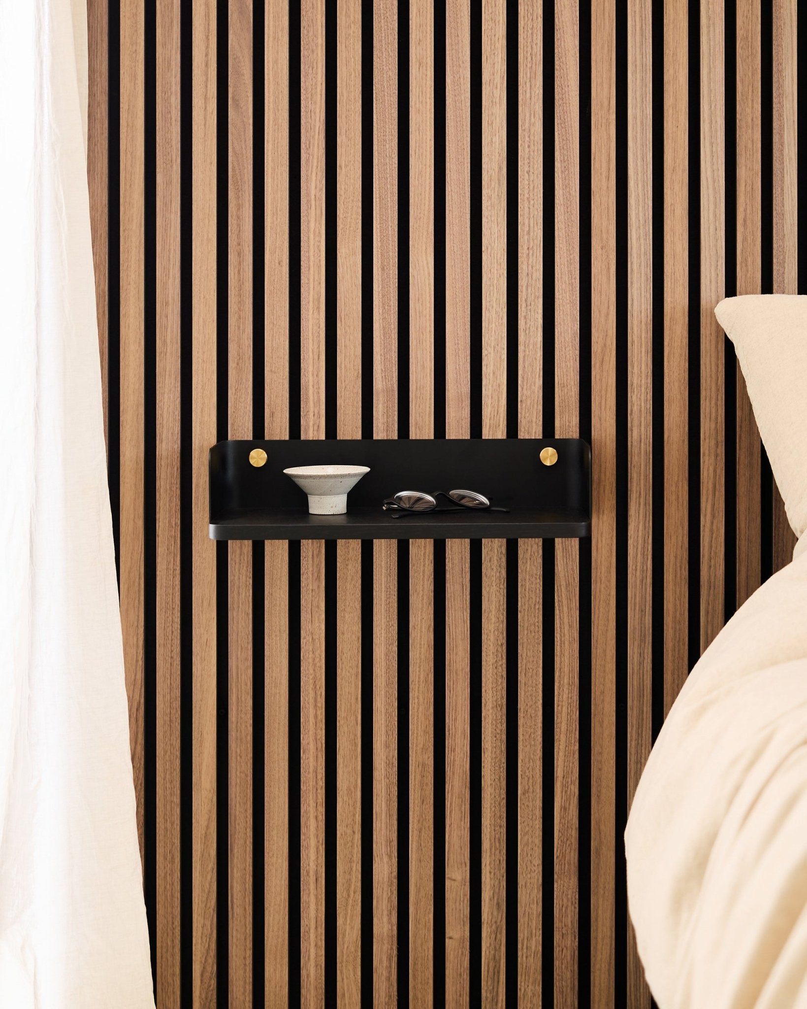 The Linn Bookshelf in Black Oak by WoodUpp is mounted on a Nordic acoustic slatwall panel, holding a bowl and glasses beside a bed with beige bedding.
