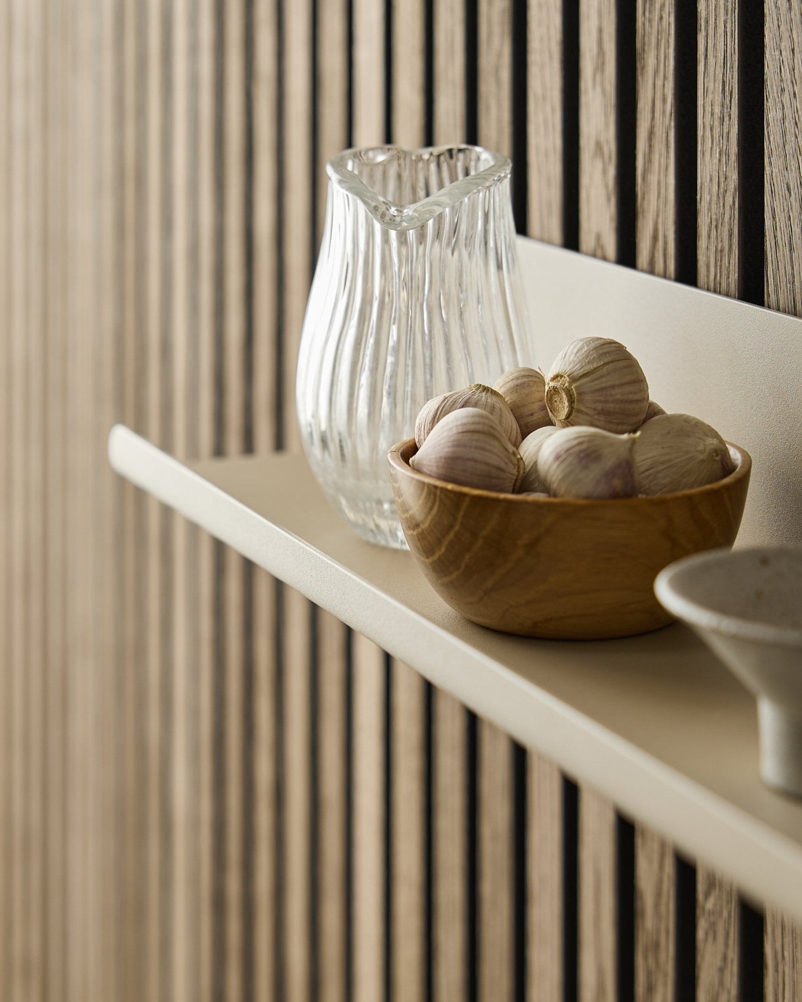 A wooden bowl of garlic and a glass jug on the Norr Wide Shelf by WoodUpp, mounted against an Akupanel acoustic panel with vertical wooden slats.