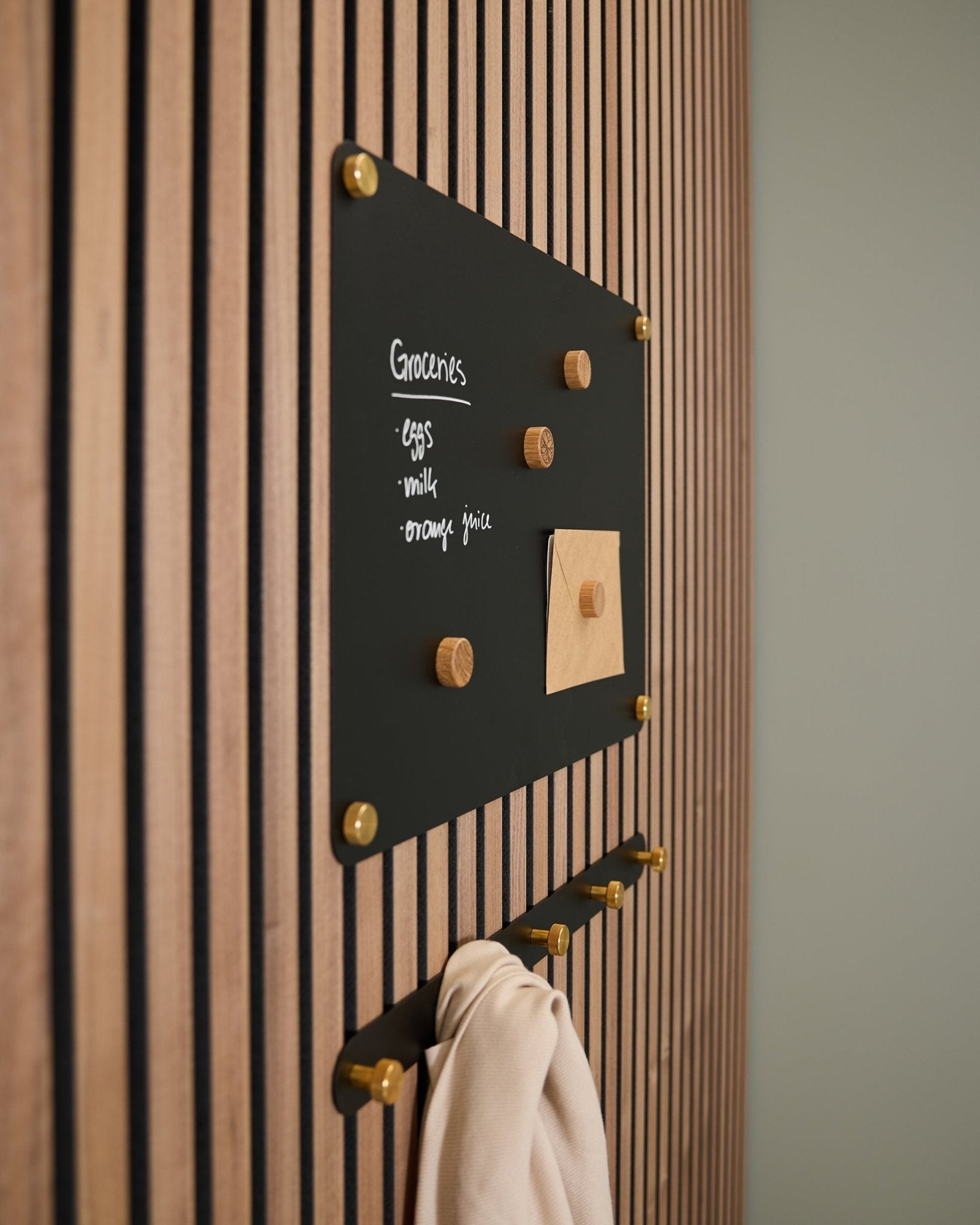 A small black Norr Magnetic Board by WoodUpp mounted on an Akupanel acoustic wood panel displays a grocery list and note, with hooks below holding a light cloth.