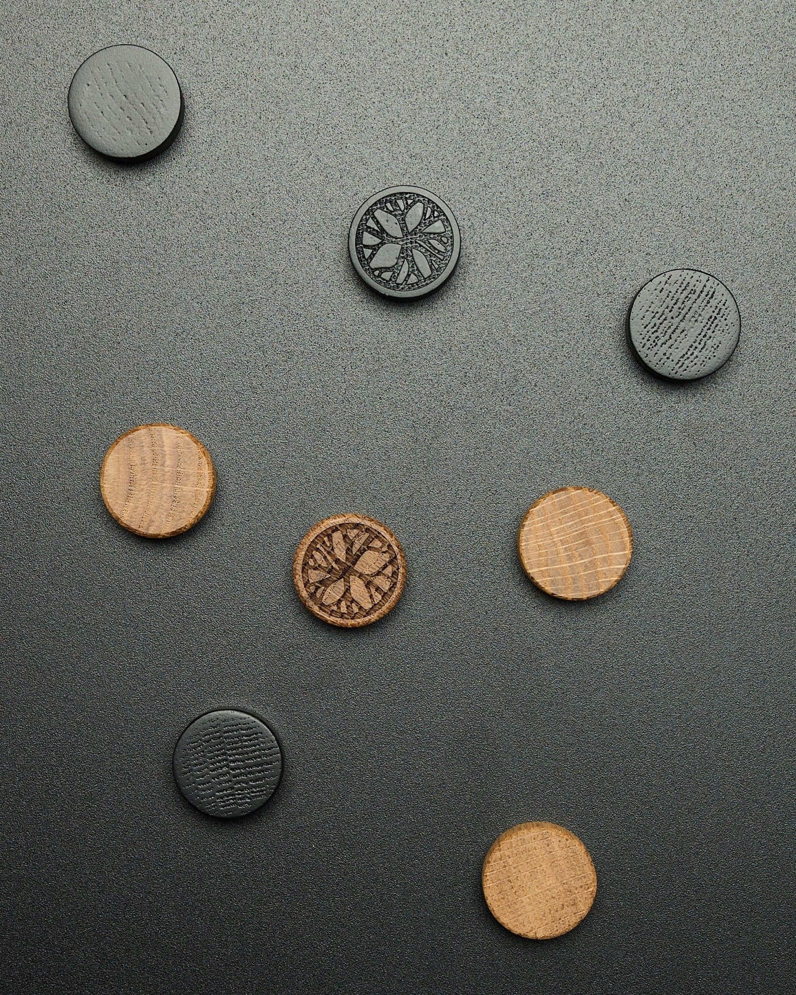 Four round black oak magnets from the WoodUpp Norr Magnet Set, loosely arranged on a textured dark surface, designed as Akupanel accessories.