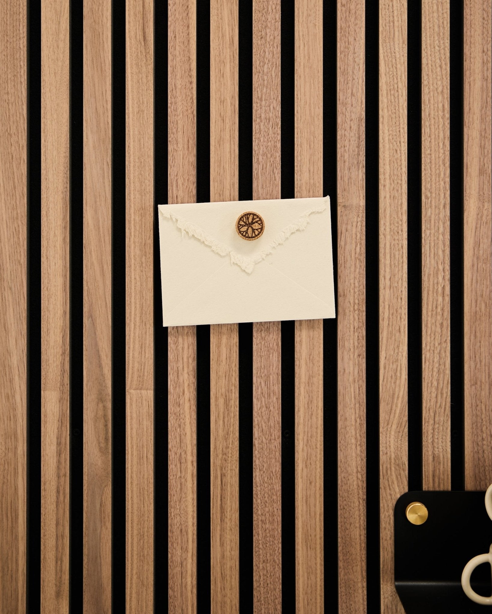 A cream envelope with a wax seal is attached to a WoodUpp oak acoustic slatwall panel using the Norr Poster Magnet set in Natural Oak; scissors and tray nearby.