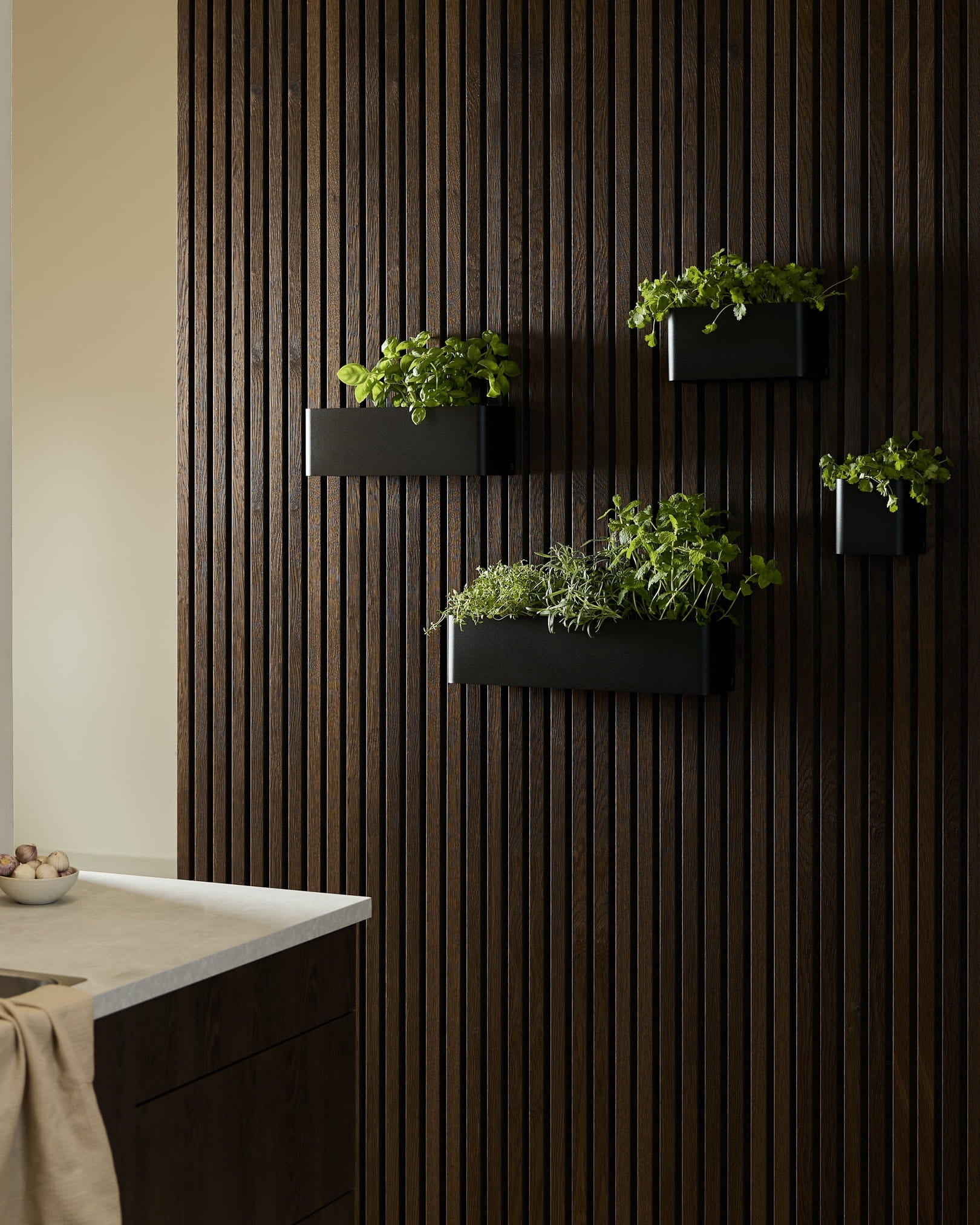 Black wall-mounted planters and a WoodUpp Norr Plant Box with green herbs on a dark wood Akupanel acoustic panel in a modern kitchen.
