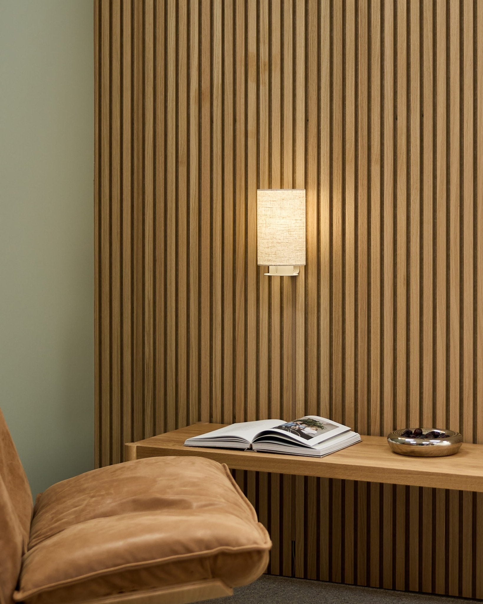 Minimal Nordic room with a tan chair, WoodUpp Akupanel acoustic slat wall, Feya Wall Lamp in Beige/Beige, an open book on a bench, and a bowl.