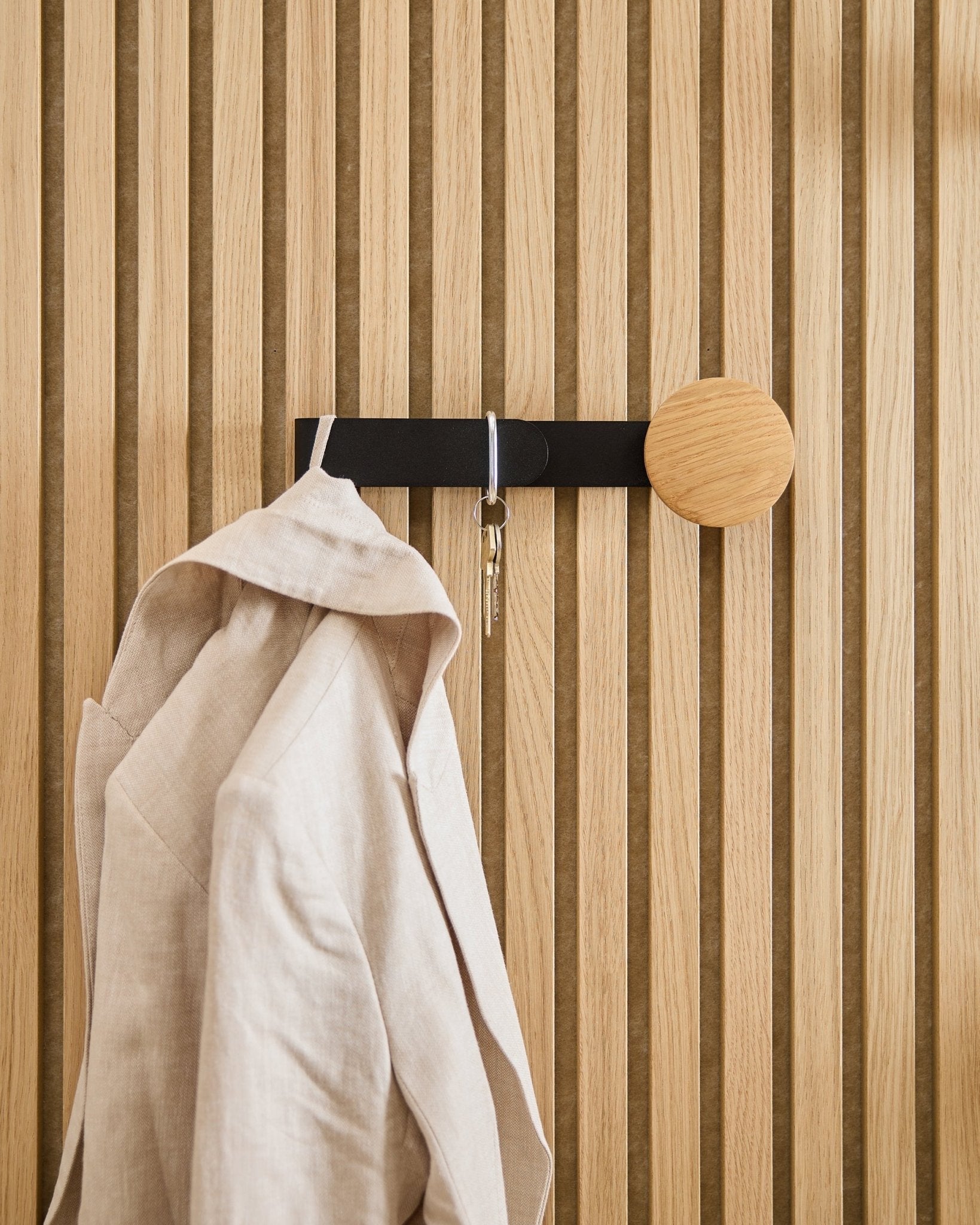 A beige blazer and a set of keys hang on the WoodUpp Linn Horizontal Hanger in natural oak, installed on a natural oak Akupanel acoustic wood panel.