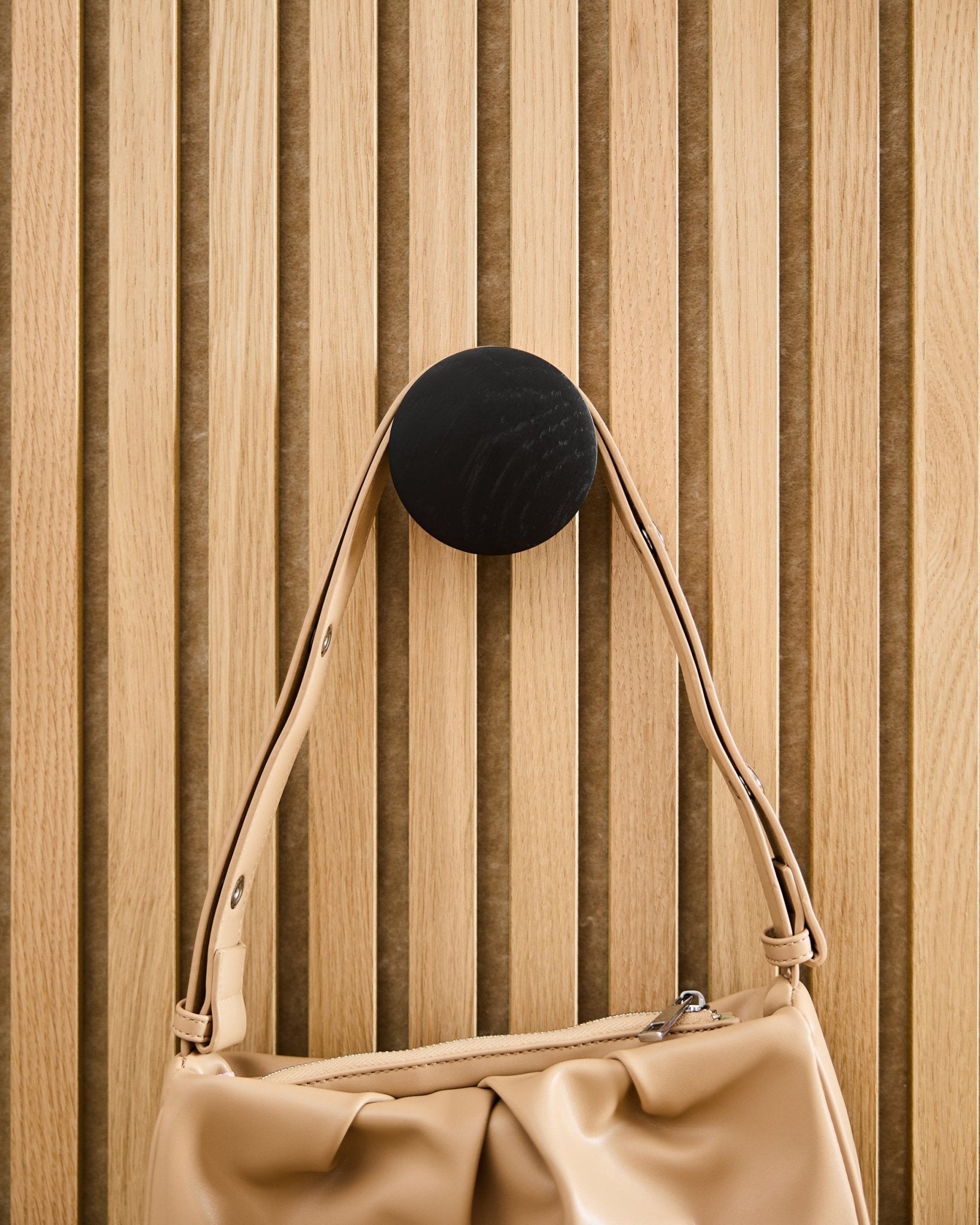 Beige handbag hanging on a WoodUpp Linn | Single Hanger in Black Oak, mounted on a light wood Akupanel acoustic slatwall panel.