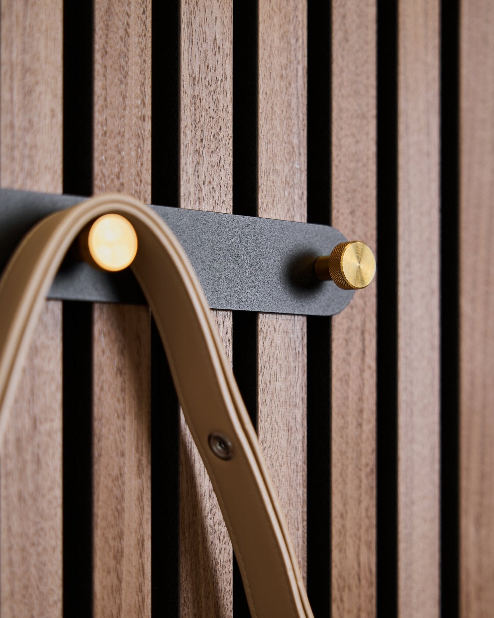 Five black/brass hooks of the Norr Coat Hook Rack by WoodUpp mounted on a wooden acoustic slatwall panel, each holding a beige strap.