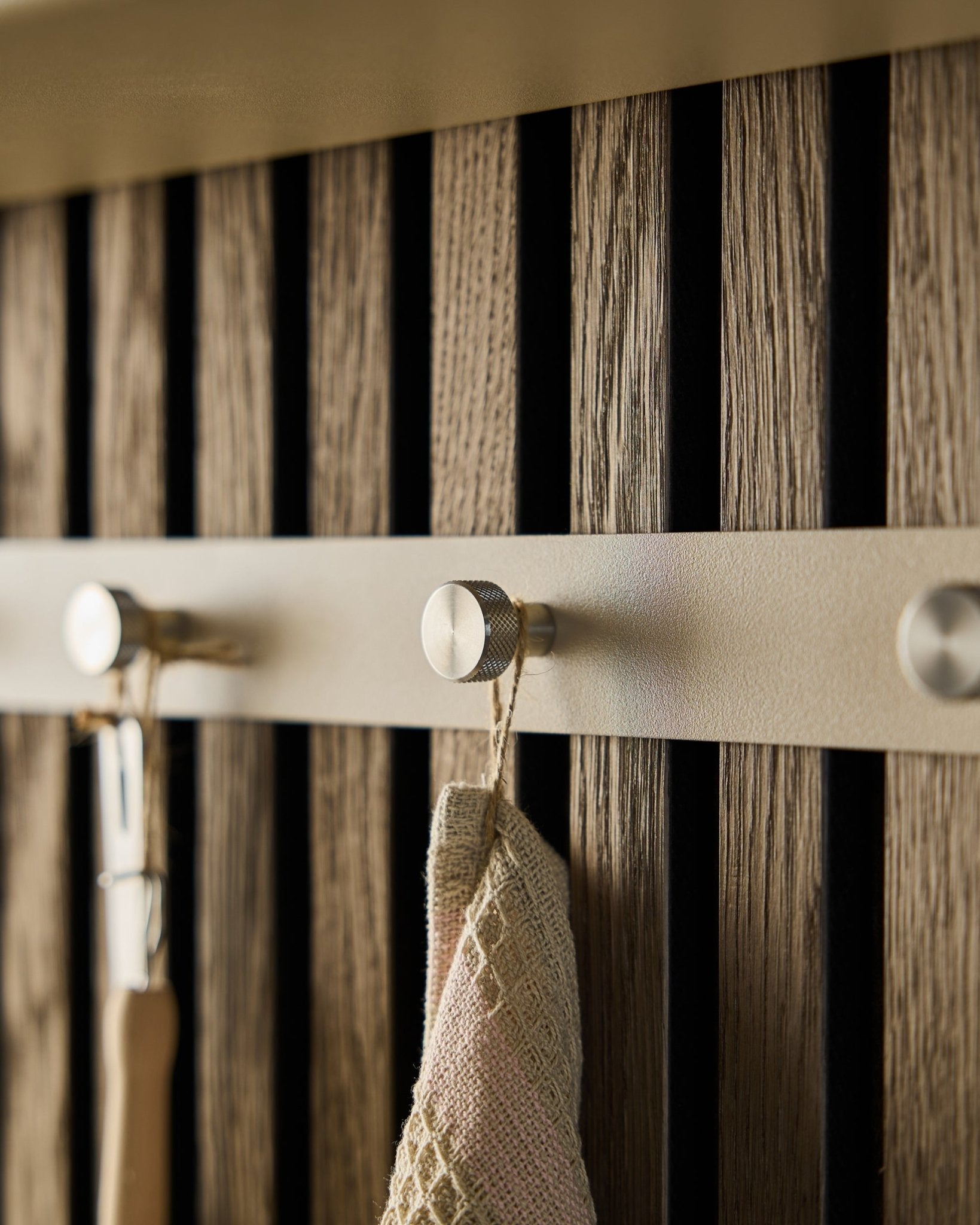 Close-up of the WoodUpp Norr Coat Hook Rack (Beige/Stainless Steel, X5) mounted on a wooden slatwall acoustic panel, holding a towel and handled tool.