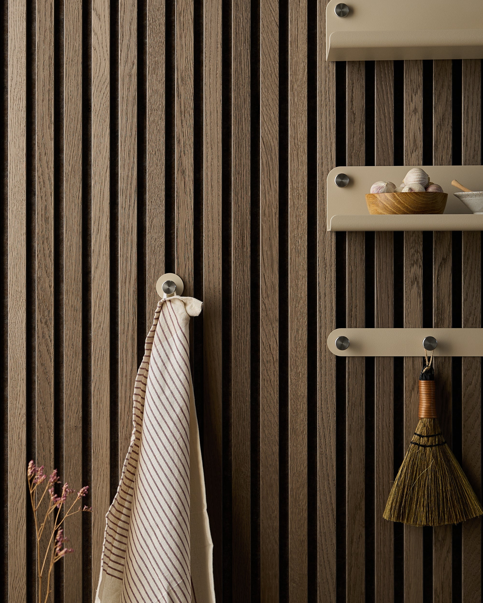 Striped towel, small broom, and bowl rest on shelves mounted to a dark Akupanel acoustic wood panel, with dried flowers at the bottom left.
