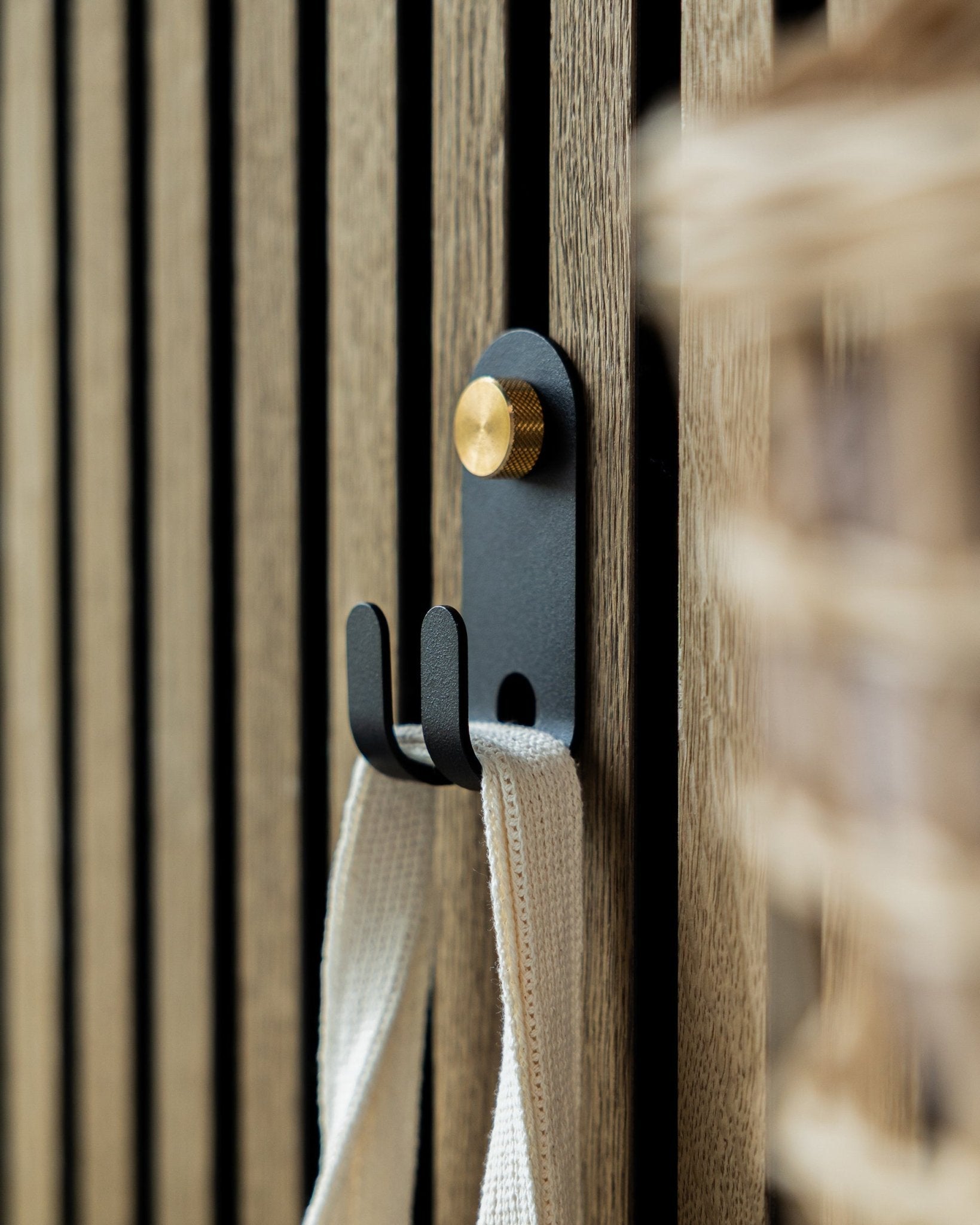 The WoodUpp Norr Double Hanger in black is mounted on a Nordic wooden acoustic slatwall panel, holding a cream-colored fabric strap.