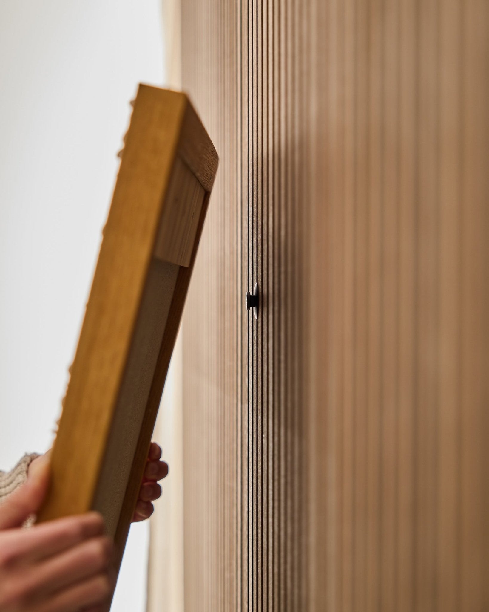 A person uses the WoodUpp Dupps | Original set to mount an Akupanel acoustic wood panel frame, focusing on attaching a small black hook near vertical slats.