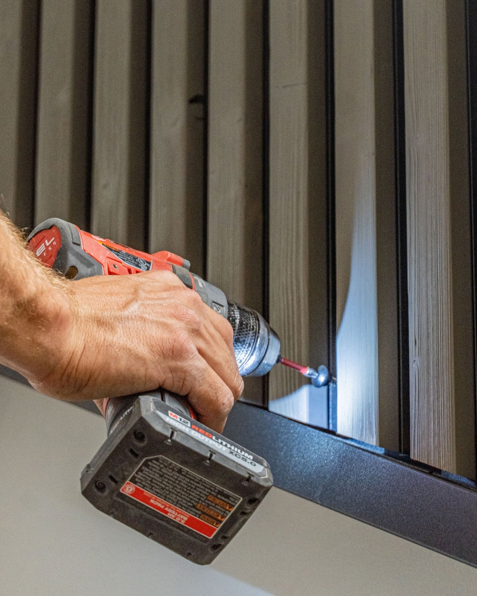 A person fastens a black WoodUpp Front Screw (4.8x35, Ø9.5 washer) into a vertical Aluwood facade panel using a cordless drill.