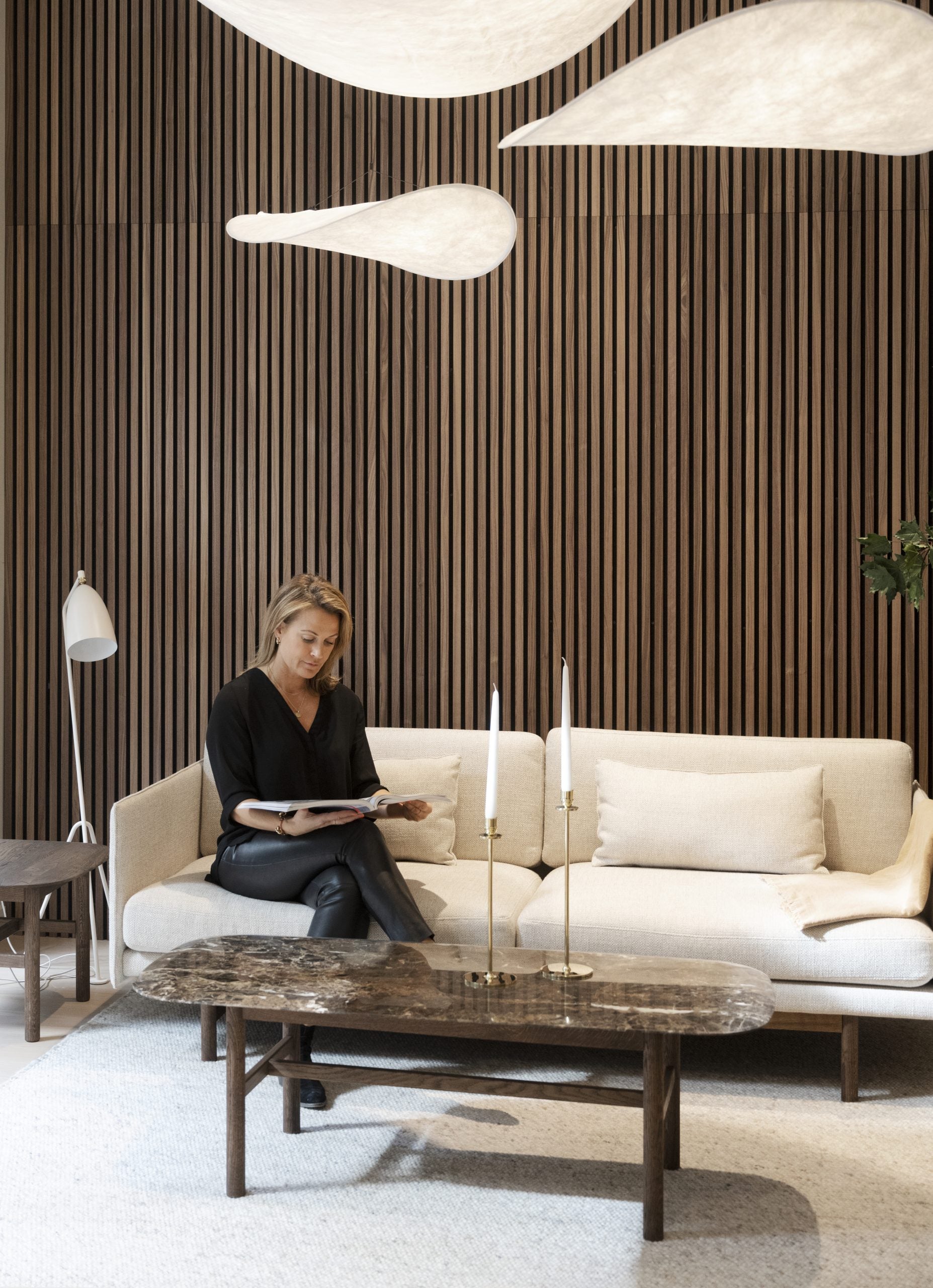 A woman reads on a cream sofa in a modern living room featuring Nordic acoustic wood panels, sculptural light fixtures, and a marble coffee table.
