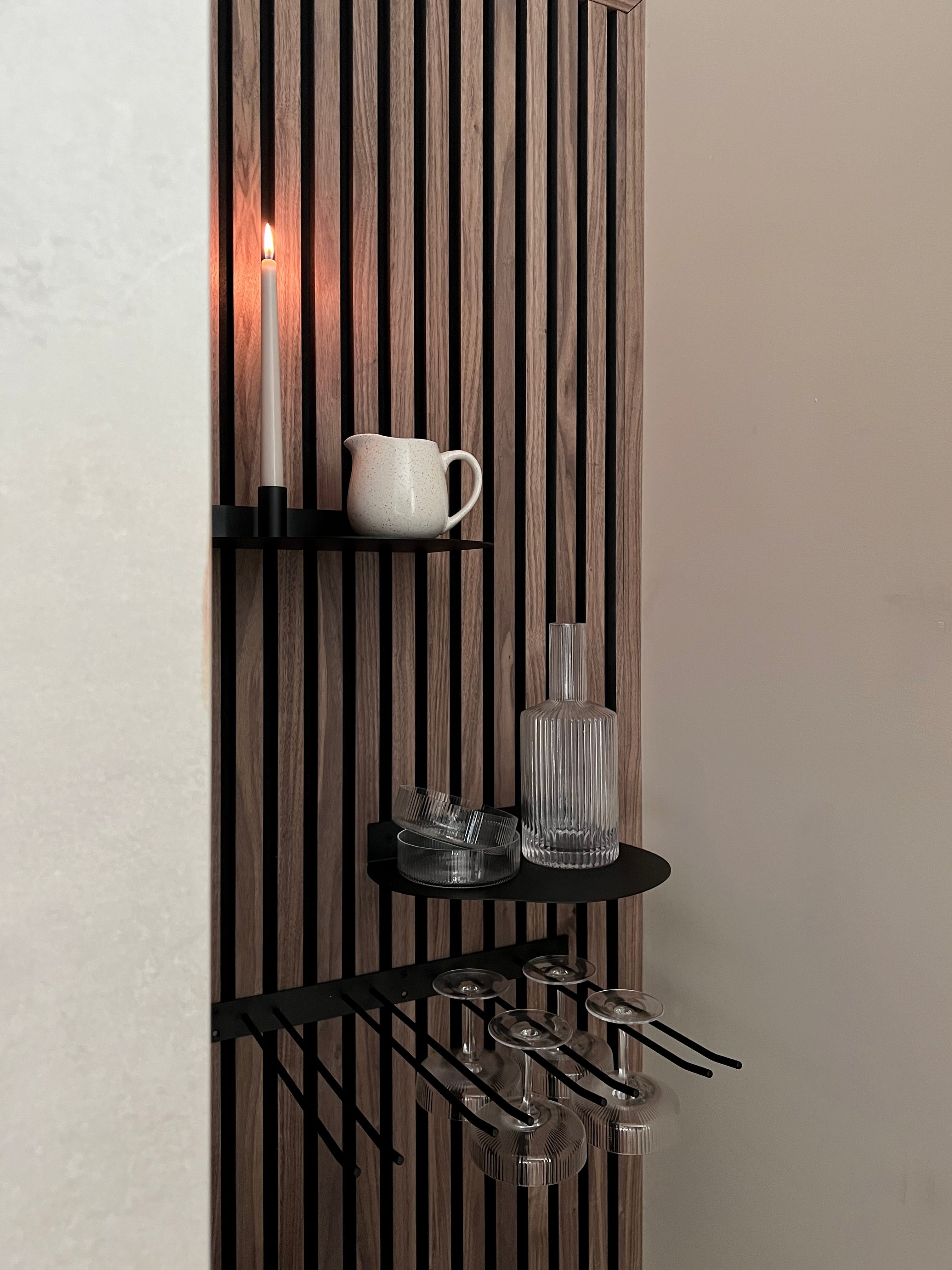 Modern Nordic acoustic slatwall panel with black shelves holding a lit candle, white pitcher, glass bottle, and stemmed wine glasses hanging below.
