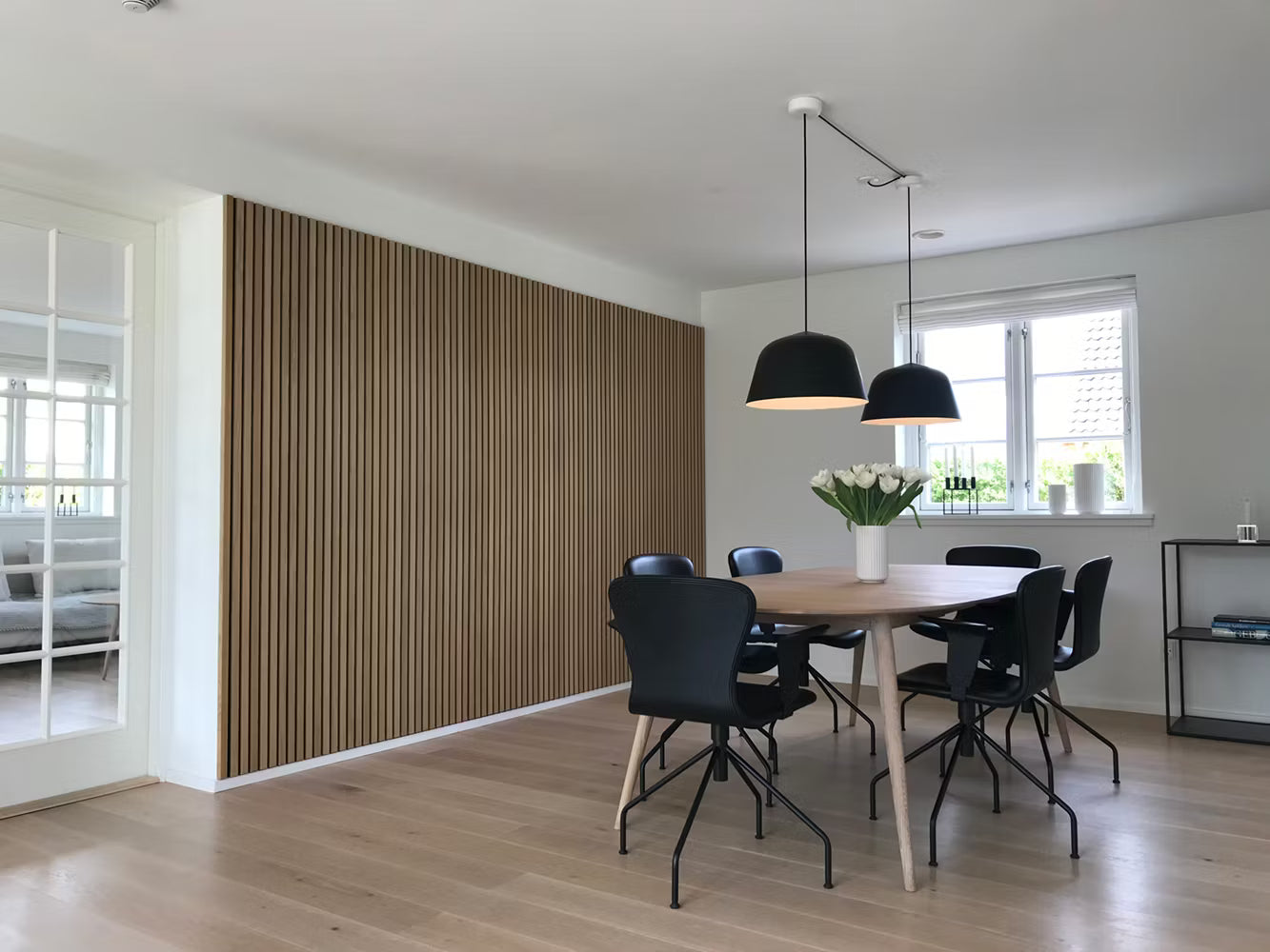 Modern dining room with wooden floor, Akupanel acoustic slatwall accent, black chairs, wooden table, pendant lights, and a white floral centerpiece.