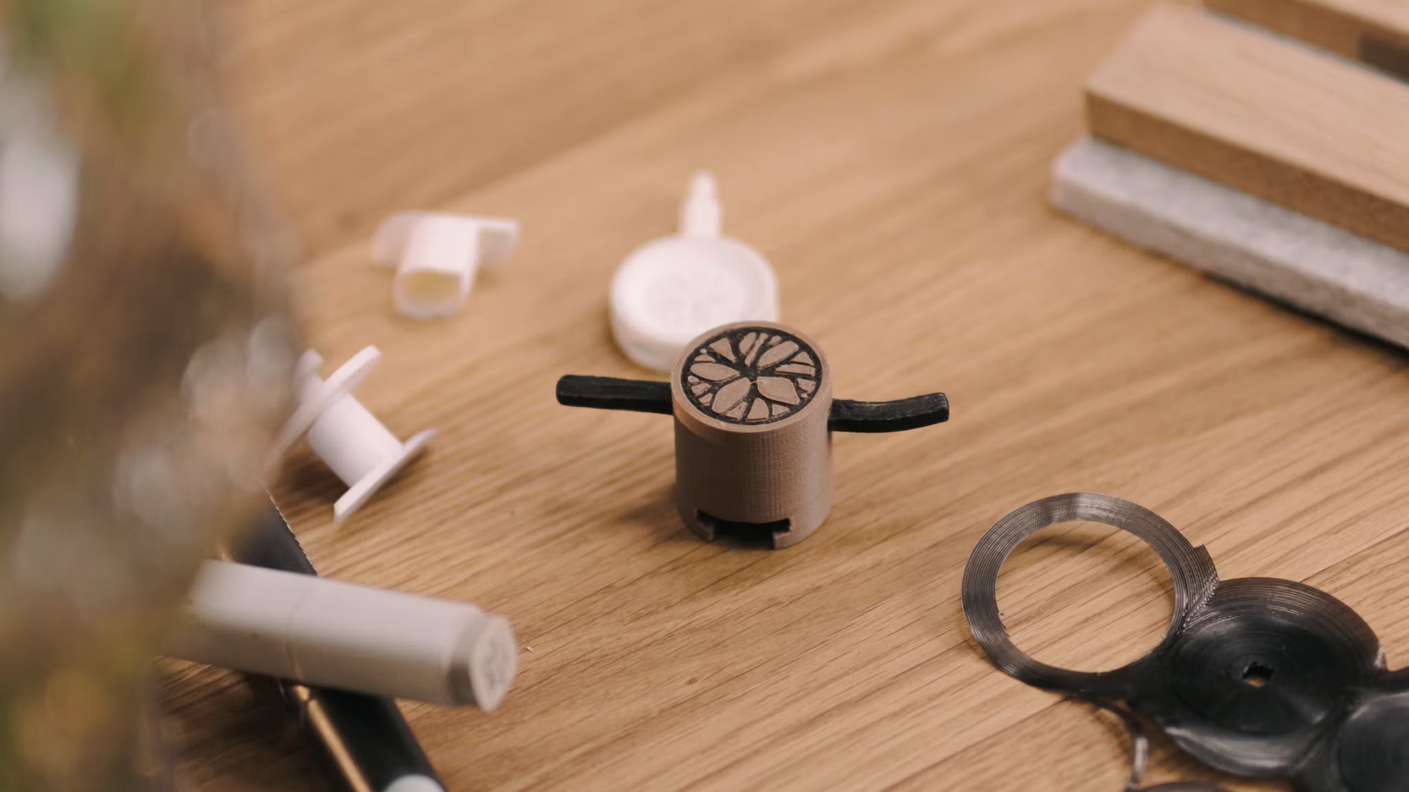 A small 3D-printed prototype with black arms sits on a wooden desk beside various plastic components, referencing acoustic panel accessory development.
