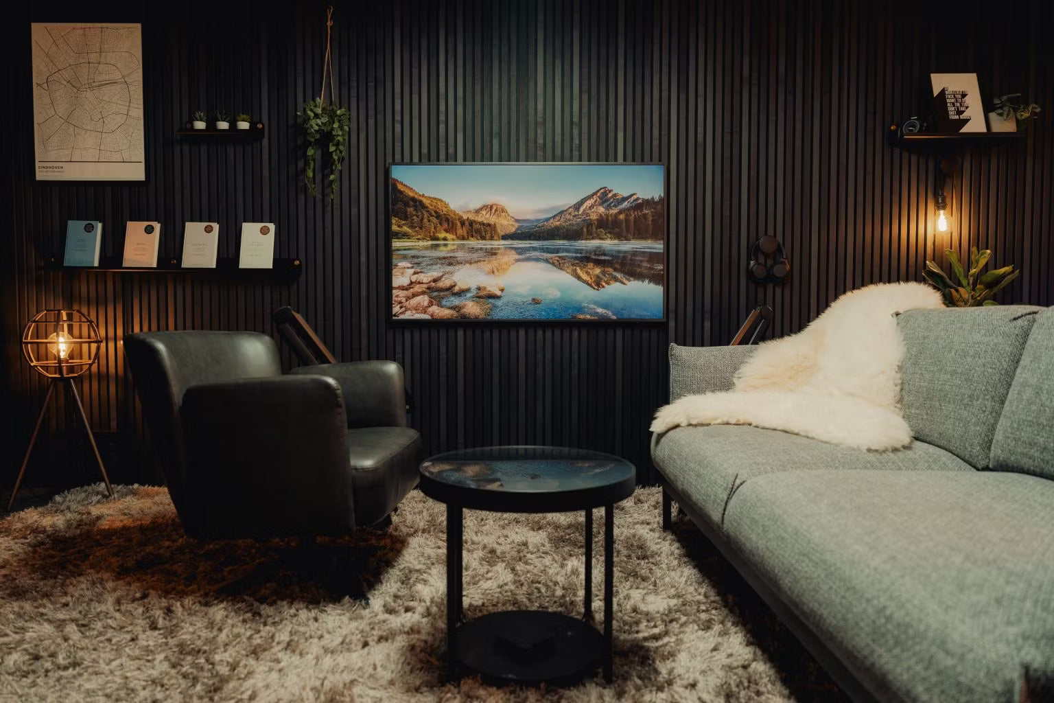A modern living room with black walls, a grey sofa, two black chairs, a round table, shaggy rug, and a landscape acoustic panel mounted on the wall.