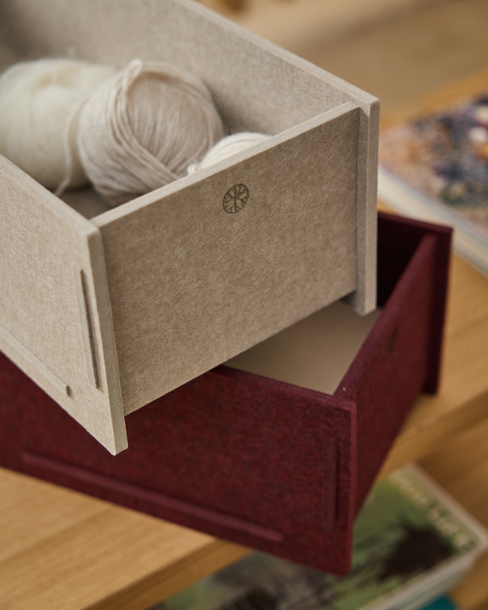 A beige PET felt storage box filled with yarn sits atop a closed WoodUpp Vigg Felt Box/BM2 in ox red, resting on wood flooring with books in the background.