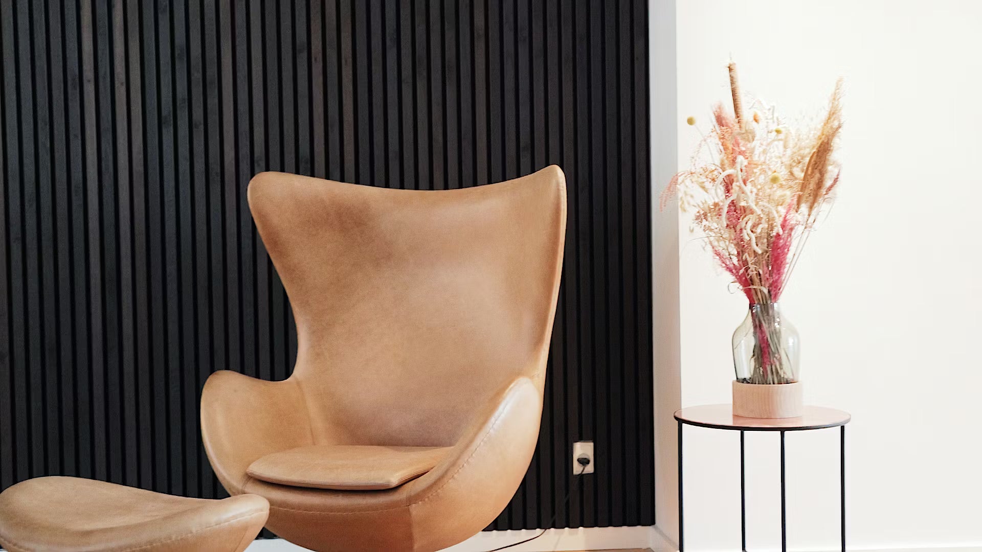 Tan leather chair and ottoman next to a small table with dried flowers, set against a black acoustic wood slat panel and white background.